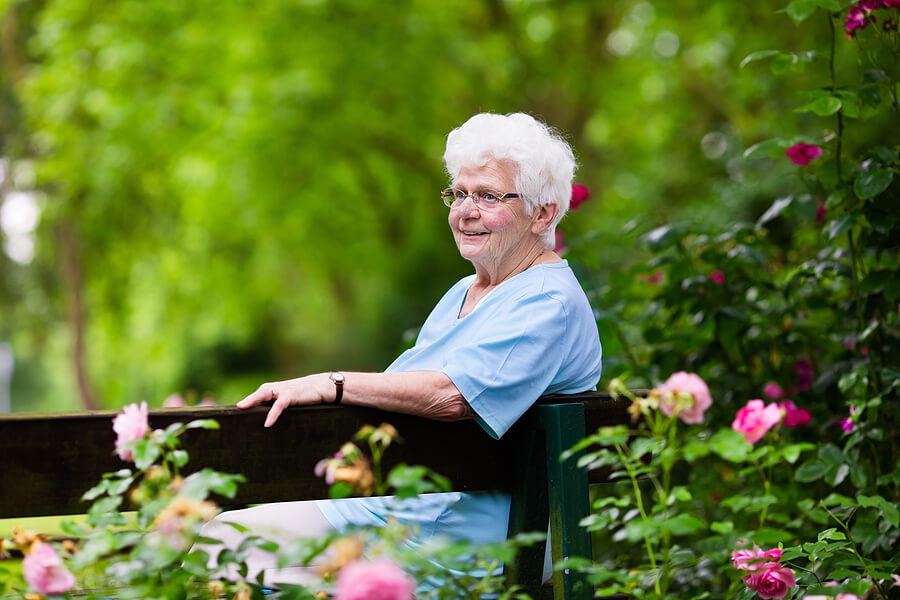 Elderly woman seated on park bench smiling and surrounded by flowers and greenery