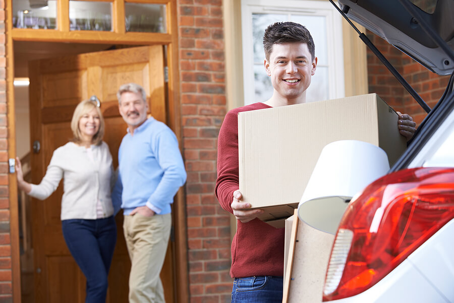 Smiling tall young adult moving boxes into back of car while his parents look on from front door smiling at him